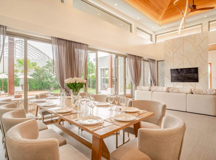 Bright dining area with a long table set for a formal meal, beige chairs, and glass doors leading outside,”“Table set for eight with plates, cutlery, wine glasses, and a vase of white tulips in the center.”,”Open living-dining space with glass walls, soft curtains, and a wall-mounted TV on a marble-patterned wall.”,”Sunny interior with high wooden ceilings, a curved chandelier, and sliding glass doors to the outdoor area.”,”Neutral, elegant dining room with large windows, outdoor greenery, and a cozy lounge area nearby.
