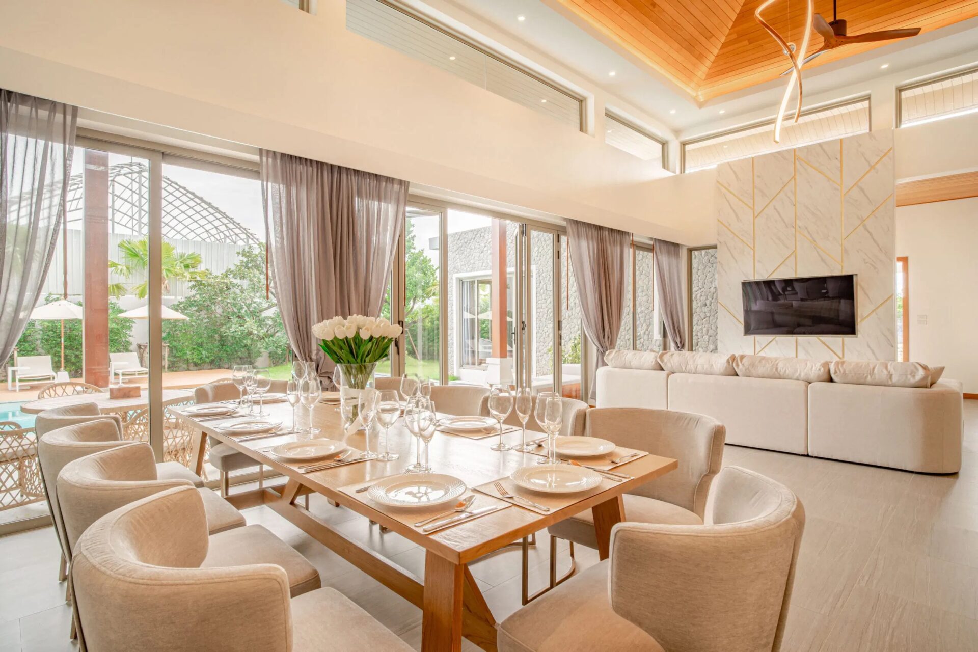 Bright dining area with a long table set for a formal meal, beige chairs, and glass doors leading outside,”“Table set for eight with plates, cutlery, wine glasses, and a vase of white tulips in the center.”,”Open living-dining space with glass walls, soft curtains, and a wall-mounted TV on a marble-patterned wall.”,”Sunny interior with high wooden ceilings, a curved chandelier, and sliding glass doors to the outdoor area.”,”Neutral, elegant dining room with large windows, outdoor greenery, and a cozy lounge area nearby.