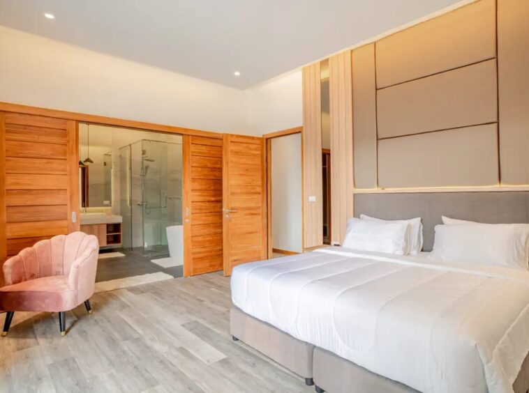 Modern hotel room with a king-size bed, white bedding, and a padded headboard set against a wood-paneled wall; a pink velvet chair sits in the corner, and a glass-enclosed bathroom with a rainfall shower is visible through sliding wooden doors in the background.