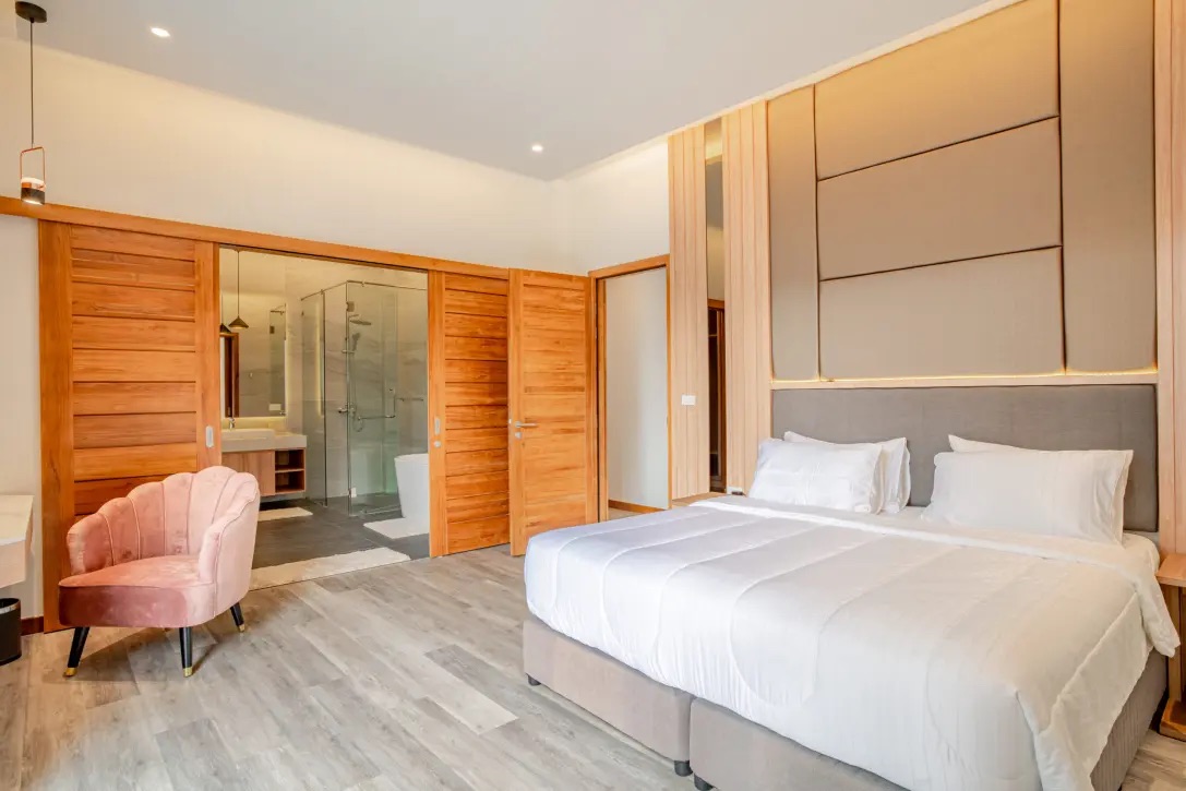 Modern hotel room with a king-size bed, white bedding, and a padded headboard set against a wood-paneled wall; a pink velvet chair sits in the corner, and a glass-enclosed bathroom with a rainfall shower is visible through sliding wooden doors in the background.