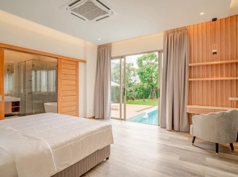 Modern hotel room with a king bed, wooden sliding doors to a glass-walled bathroom, and a view of a pool through large sliding doors on the patio.