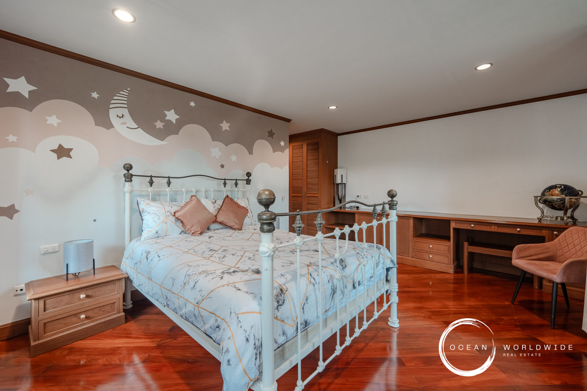 Bedroom with a pastel mural of moons and stars behind a white iron bed, marble-patterned bedding, wooden floor, and built-in desk area. Watermark reads 'Ocean Worldwide Real Estate' in the corner.