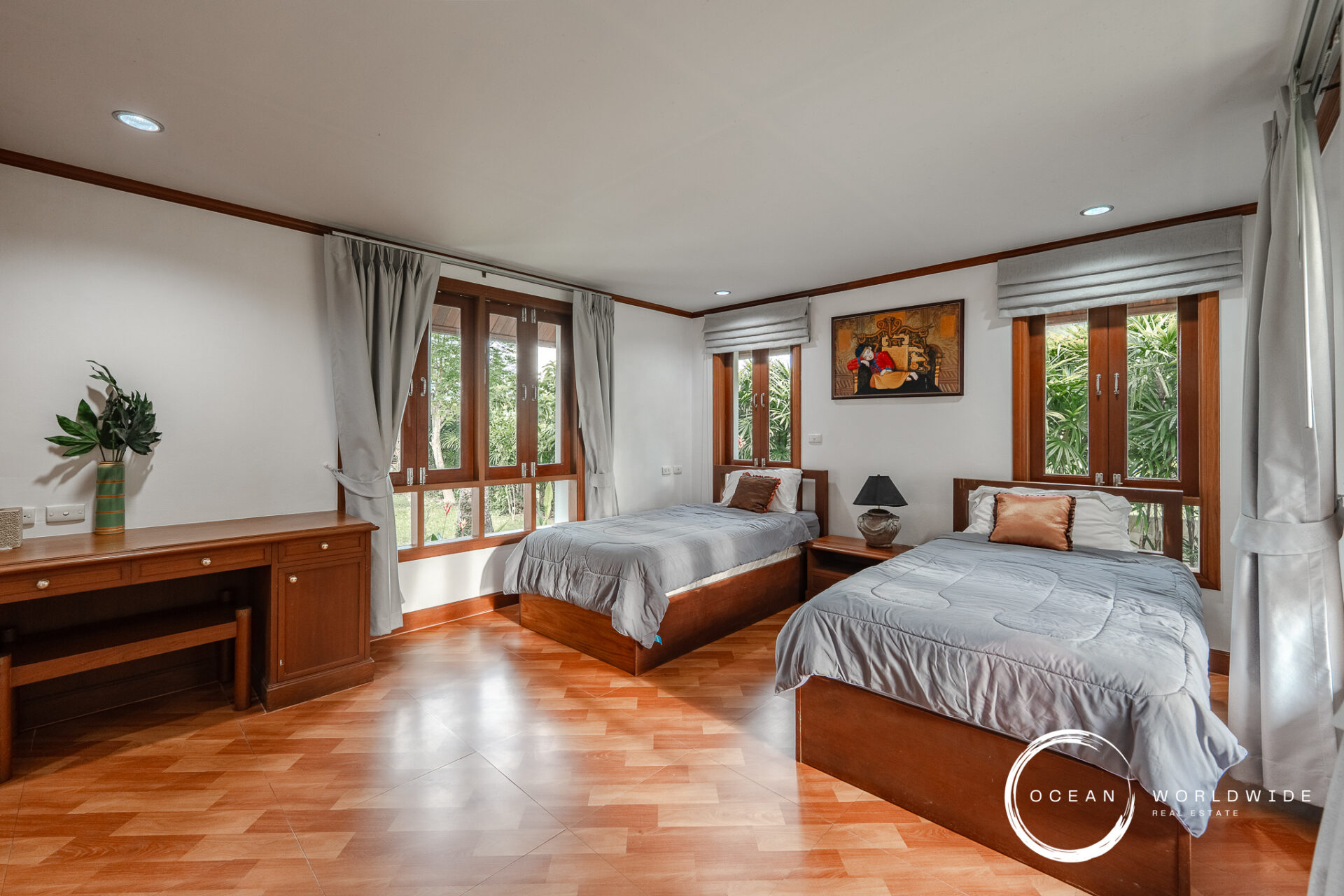 Twin beds in a bright wood-furnished bedroom with large windows, gray bedding, and a framed painting above the headboard.