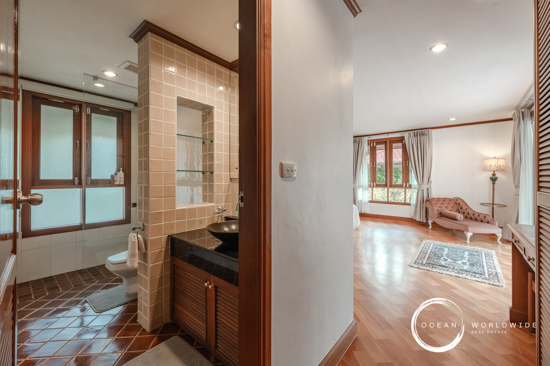 Bathroom with beige tiled walls and glass shower enclosure, next to a dark wood vanity and toilet, open to a living room view.