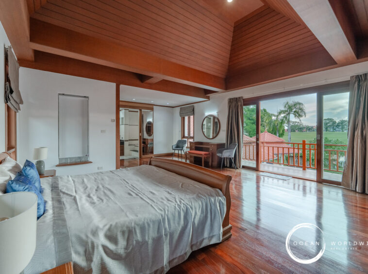 King-size bed in a bright suite with a vaulted wooden ceiling, large glass doors opening to a balcony with a lawn and palm trees outside