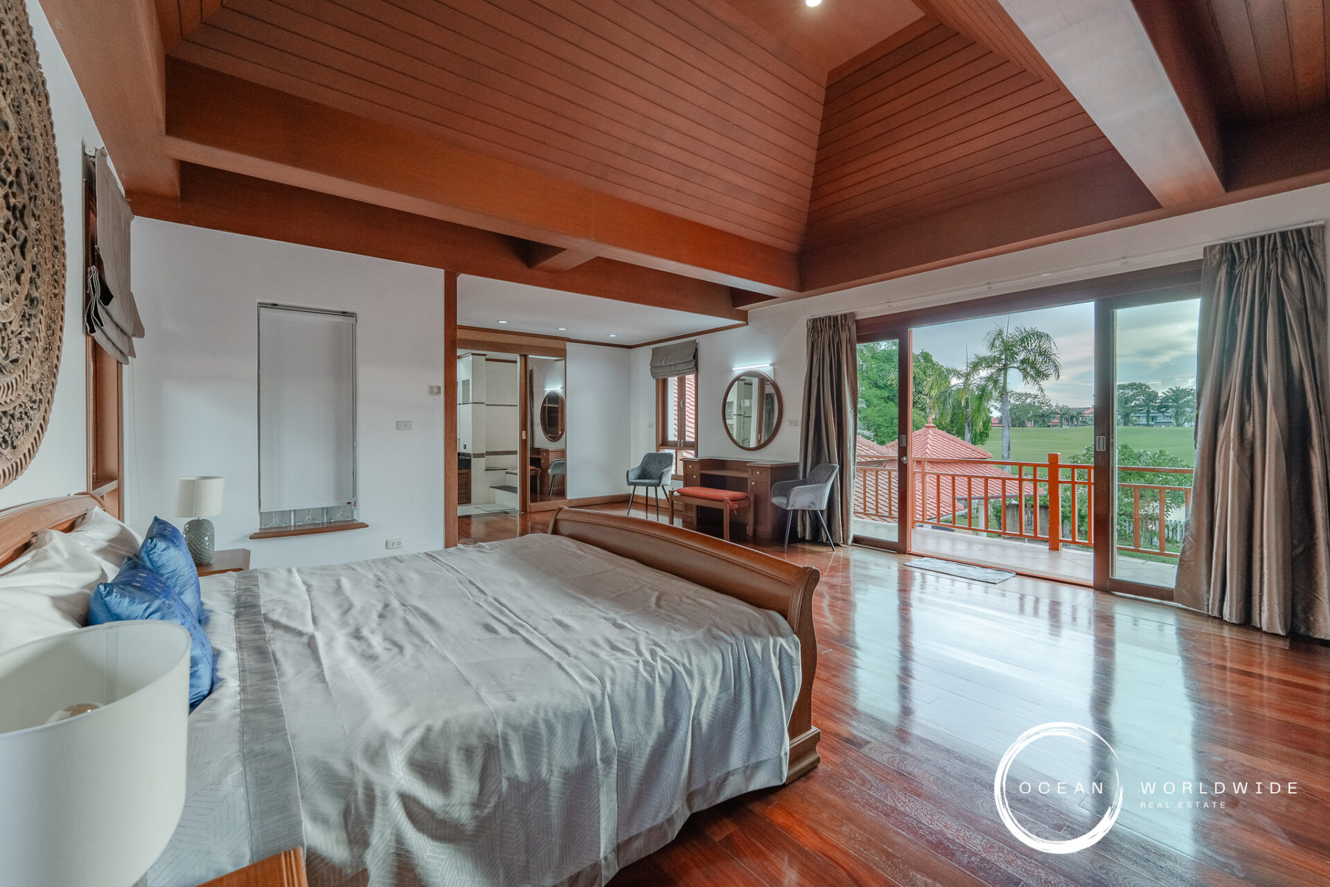 King-size bed in a bright suite with a vaulted wooden ceiling, large glass doors opening to a balcony with a lawn and palm trees outside