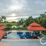 Resort pool area with red-tiled roofs, palm trees, and a green lawn beyond a water feature under a cloudy sky, logo in corner