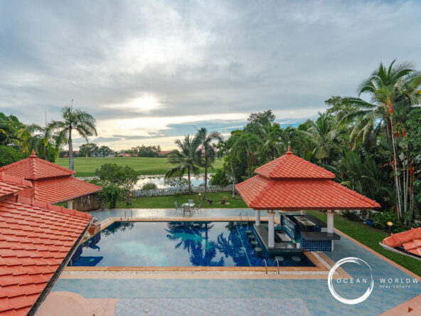Resort pool area with red-tiled roofs, palm trees, and a green lawn beyond a water feature under a cloudy sky, logo in corner