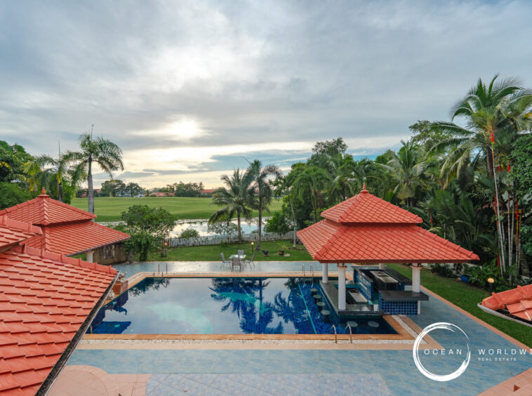 Resort pool area with red-tiled roofs, palm trees, and a green lawn beyond a water feature under a cloudy sky, logo in corner