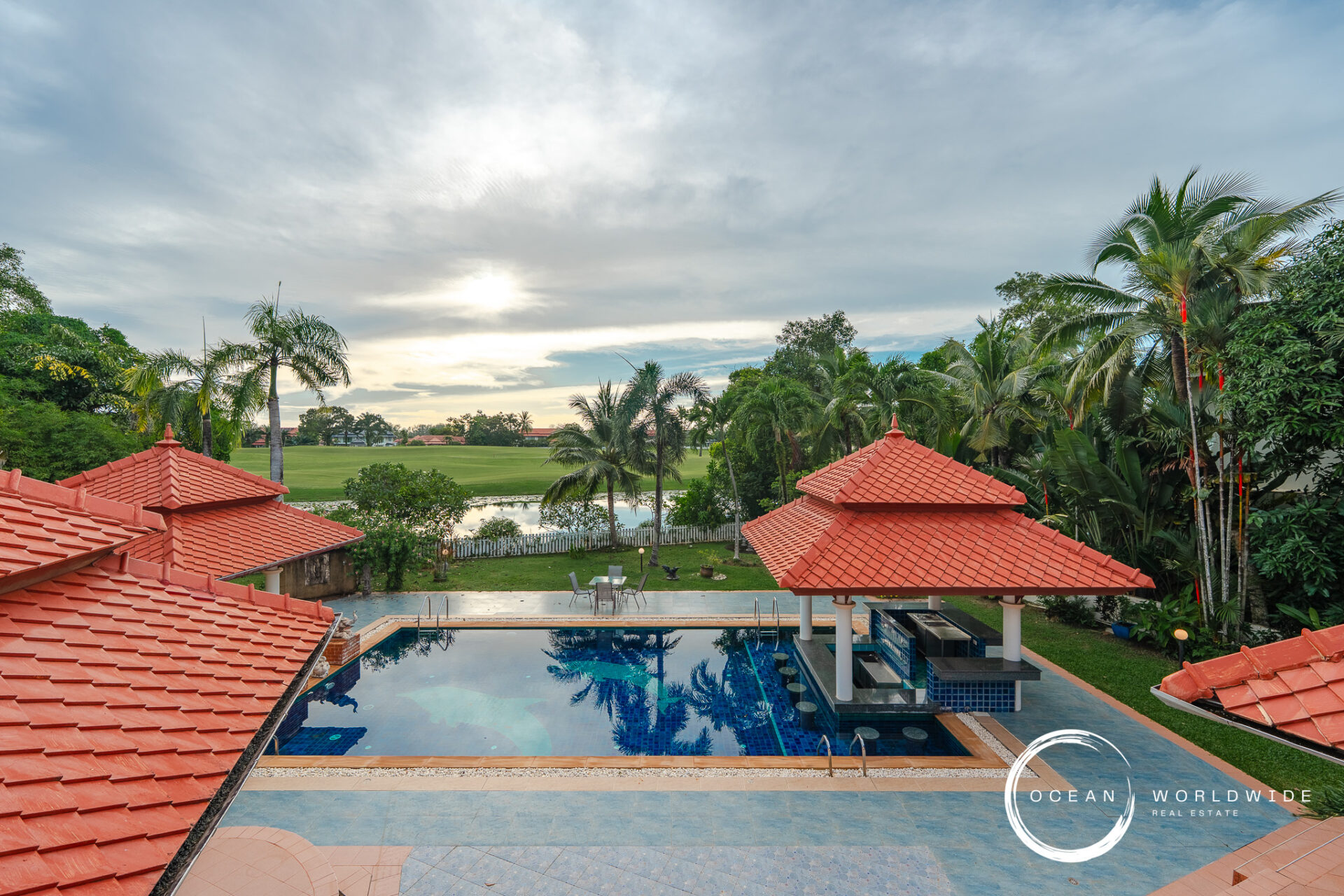Resort pool area with red-tiled roofs, palm trees, and a green lawn beyond a water feature under a cloudy sky, logo in corner