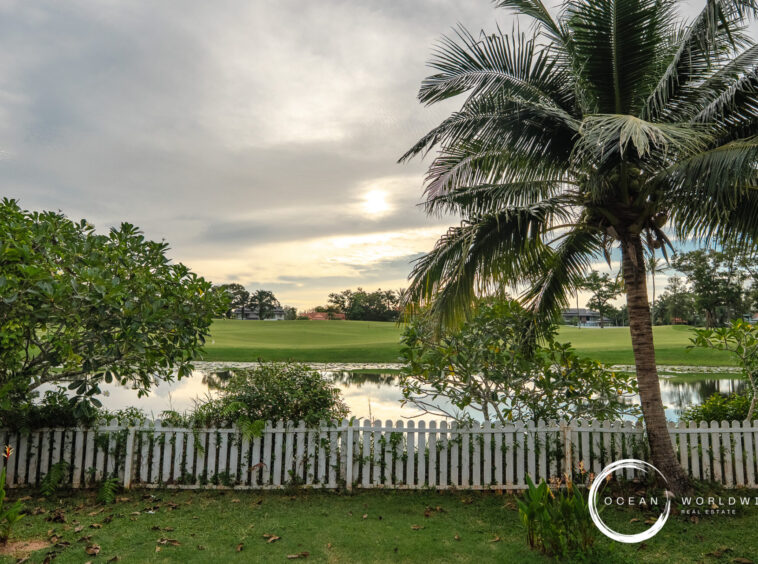 Backyard with a white picket fence, a tall palm, dense shrubs, and a reflective pond over green lawn.
