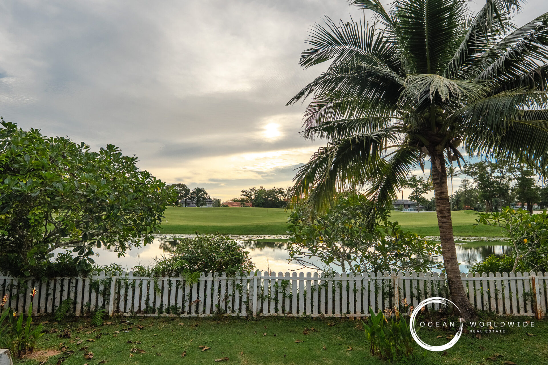Backyard with a white picket fence, a tall palm, dense shrubs, and a reflective pond over green lawn.