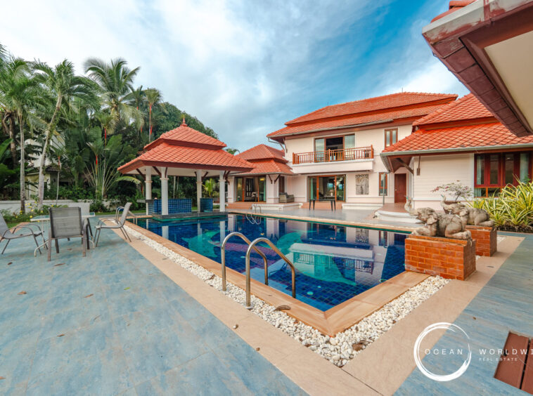 Luxurious tropical villa with red-tiled roofs, a manicured garden, and a rectangular blue pool surrounded by lounge chairs.