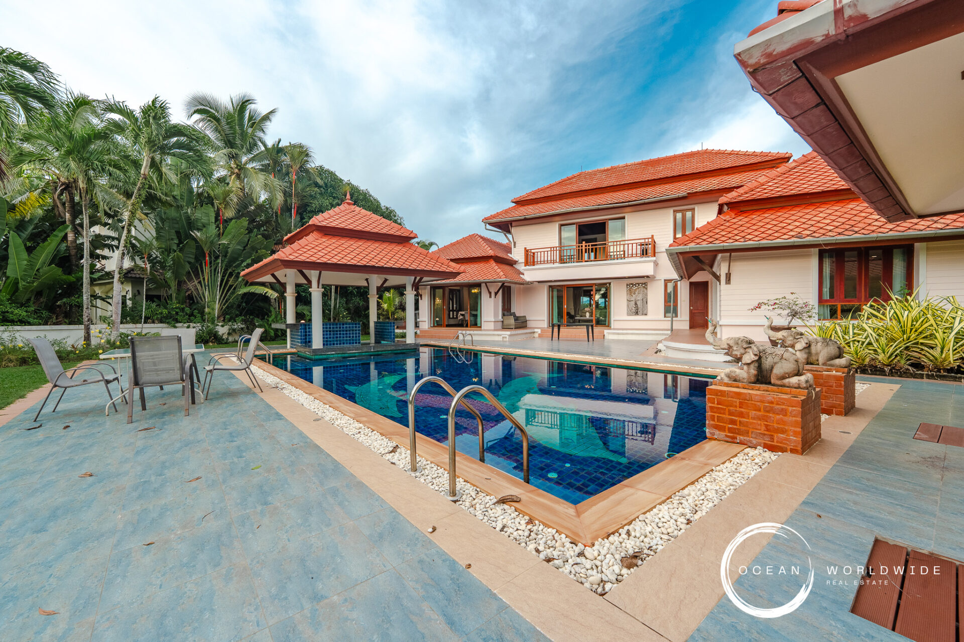 Luxurious tropical villa with red-tiled roofs, a manicured garden, and a rectangular blue pool surrounded by lounge chairs.