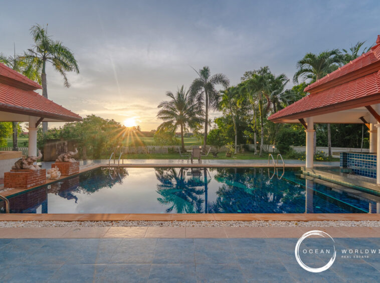 Sunset over a resort pool with palm trees and red-tiled pavilions, reflected in the water.