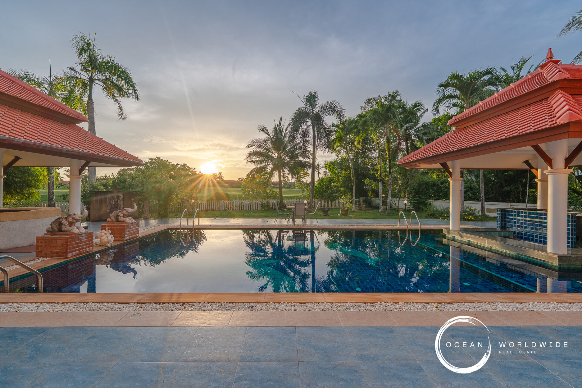 Sunset over a resort pool with palm trees and red-tiled pavilions, reflected in the water.