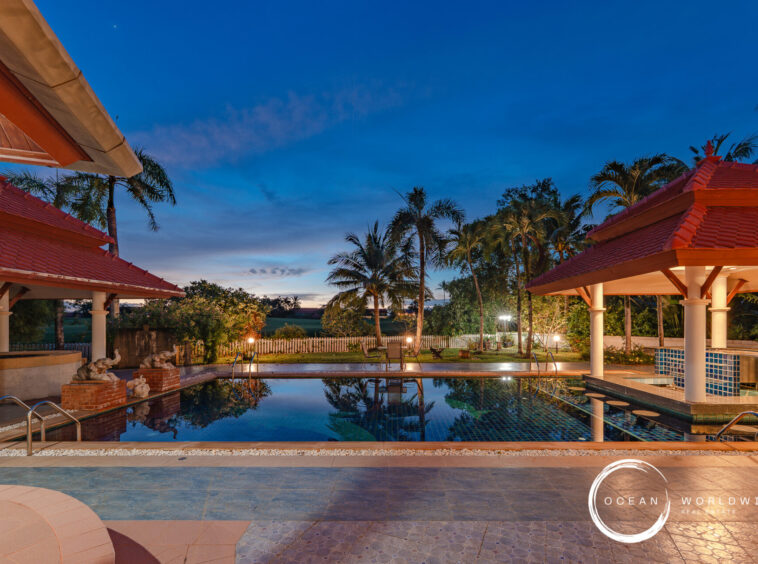 Resort-style pool area at dusk with red-tiled pavilion roofs, white columns, and palm trees framing a calm reflecting pool