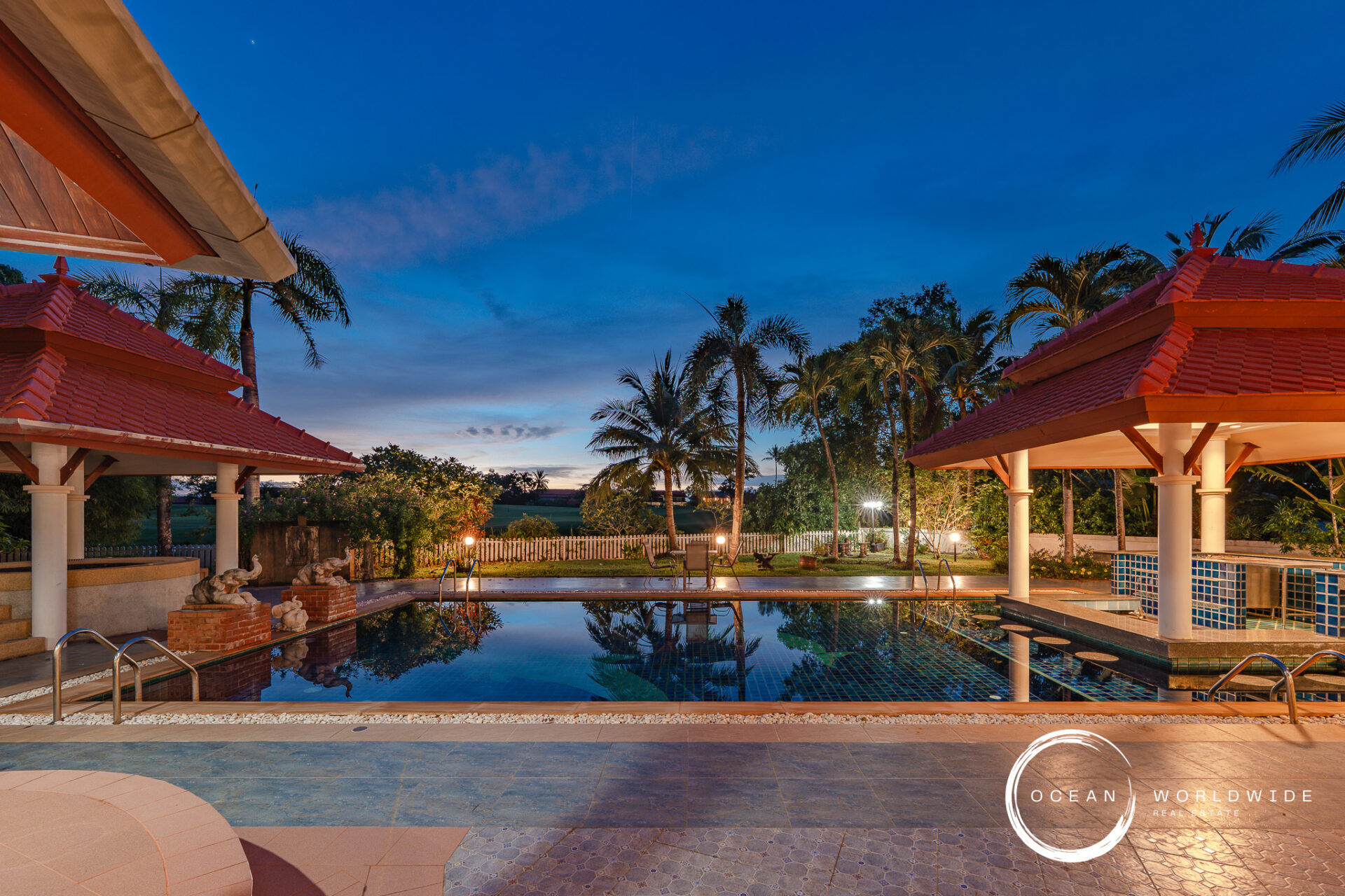 Resort-style pool area at dusk with red-tiled pavilion roofs, white columns, and palm trees framing a calm reflecting pool