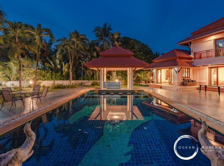 Luxurious backyard pool area with a gazebo, palm trees, and a red-tiled, two-story villa at dusk