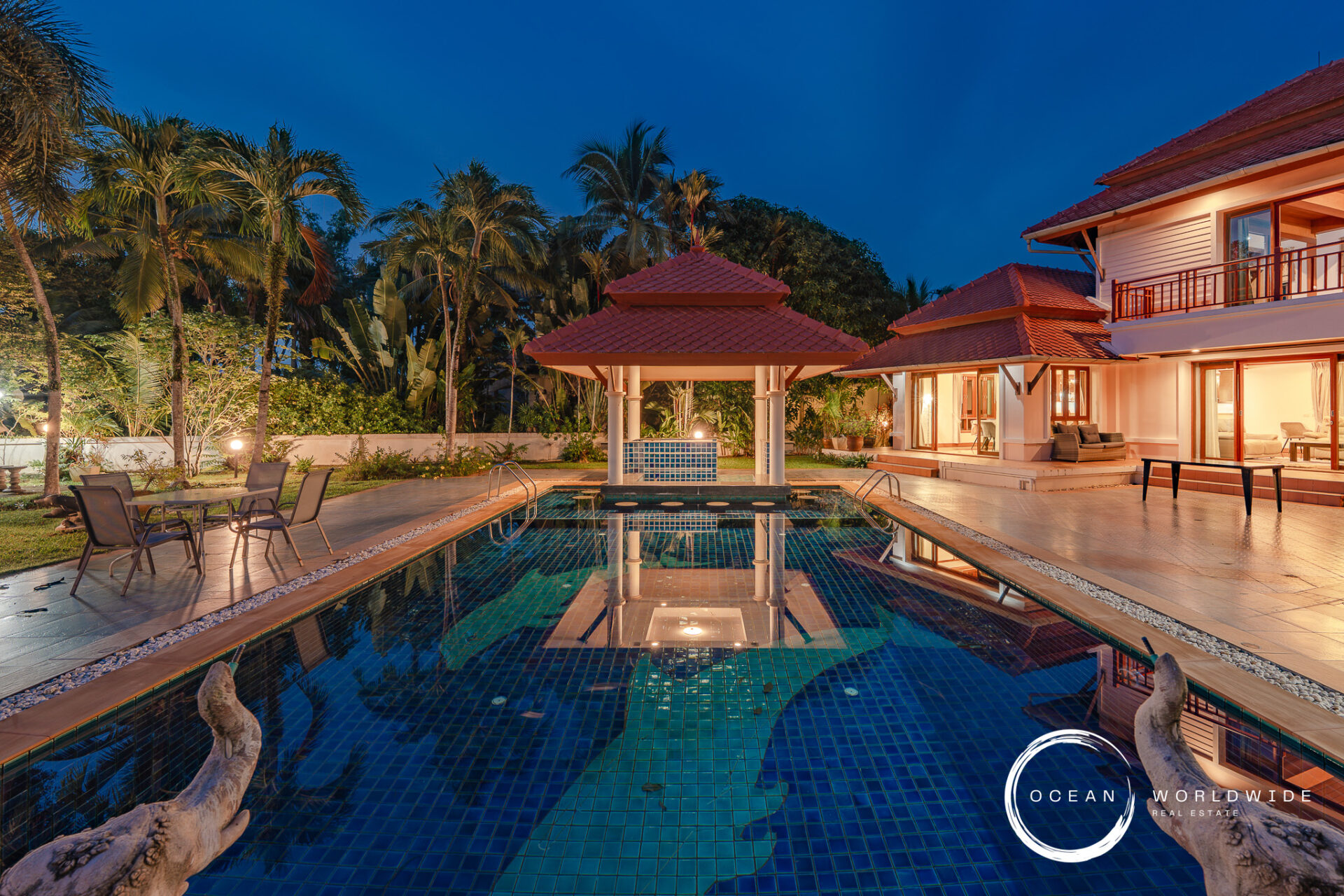 Luxurious backyard pool area with a gazebo, palm trees, and a red-tiled, two-story villa at dusk