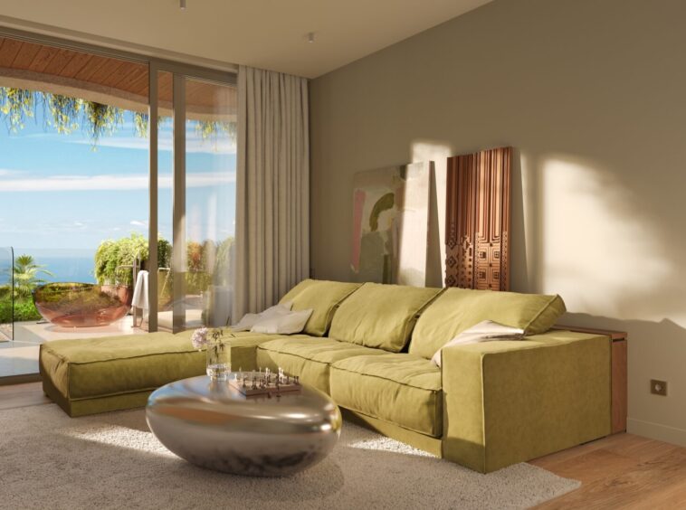 Living room with a green sectional sofa, glass doors to a sunny balcony, and ocean view beyond plants outside.