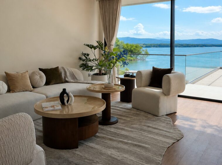 Modern living room with cream sofa and round marble coffee tables, plush throw pillows, and potted plants, open to a glass wall overlooking a lake and distant mountains.