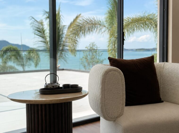 Cozy living room with a textured beige chair, brown cushion, and a round marble table by a glass wall overlooking palm trees and a blue ocean.