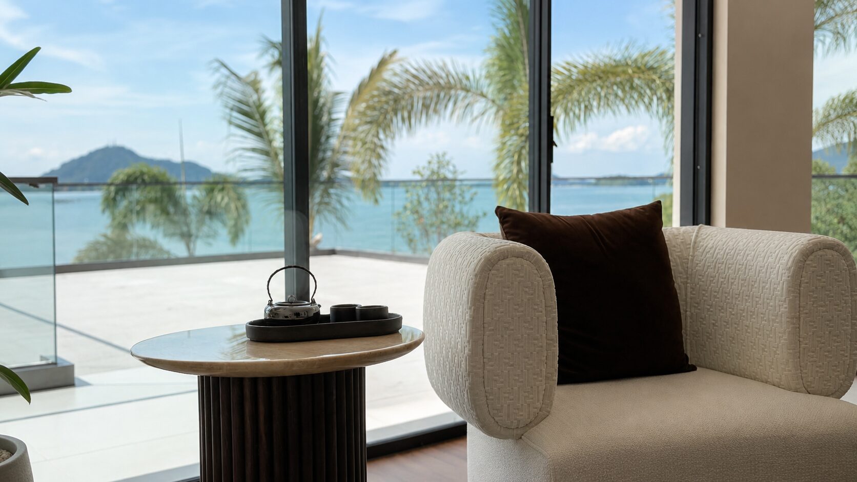 Cozy living room with a textured beige chair, brown cushion, and a round marble table by a glass wall overlooking palm trees and a blue ocean.