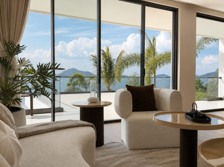 Bright living room with beige sofas, round glass-top tables, and floor-to-ceiling windows overlooking the sea and palm trees outside.
