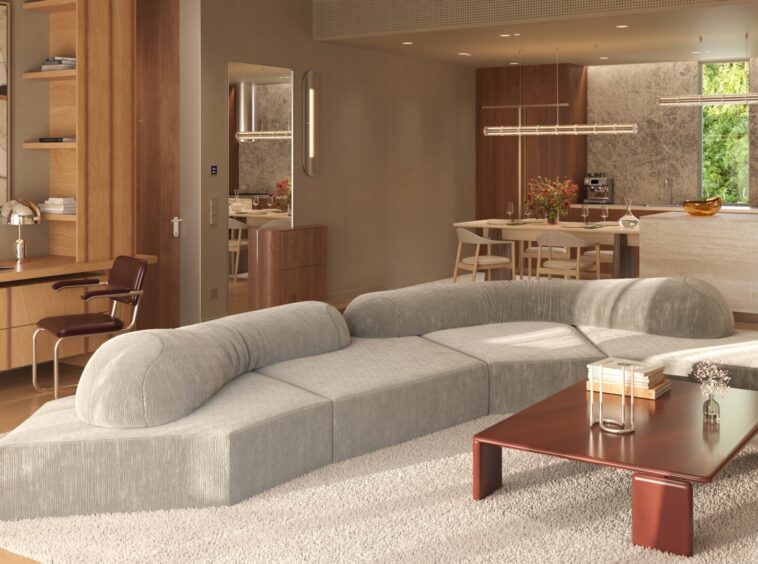 Open-concept living room with a large gray sectional on a white shag rug, a wooden coffee table, a desk and shelves, and a warm wood-toned kitchen and dining area in the background.