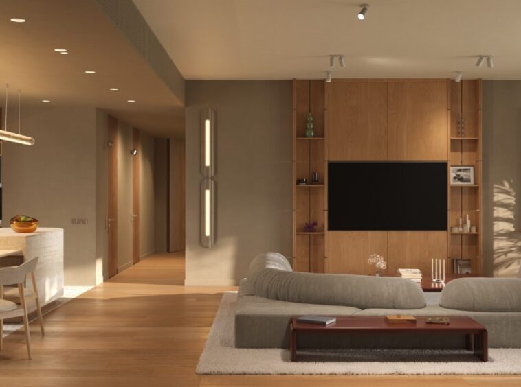 Open-plan living area with a wooden kitchen, round dining table, and a gray sectional facing a wall-mounted TV in a warm, modern apartment, with a balcony seen through large glass doors.