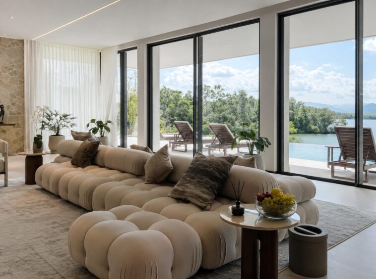 Luxurious living room with a cream tufted sectional, neutral throw pillows, and a stone accent wall, opening to a terrace with a lake view through large glass doors.
