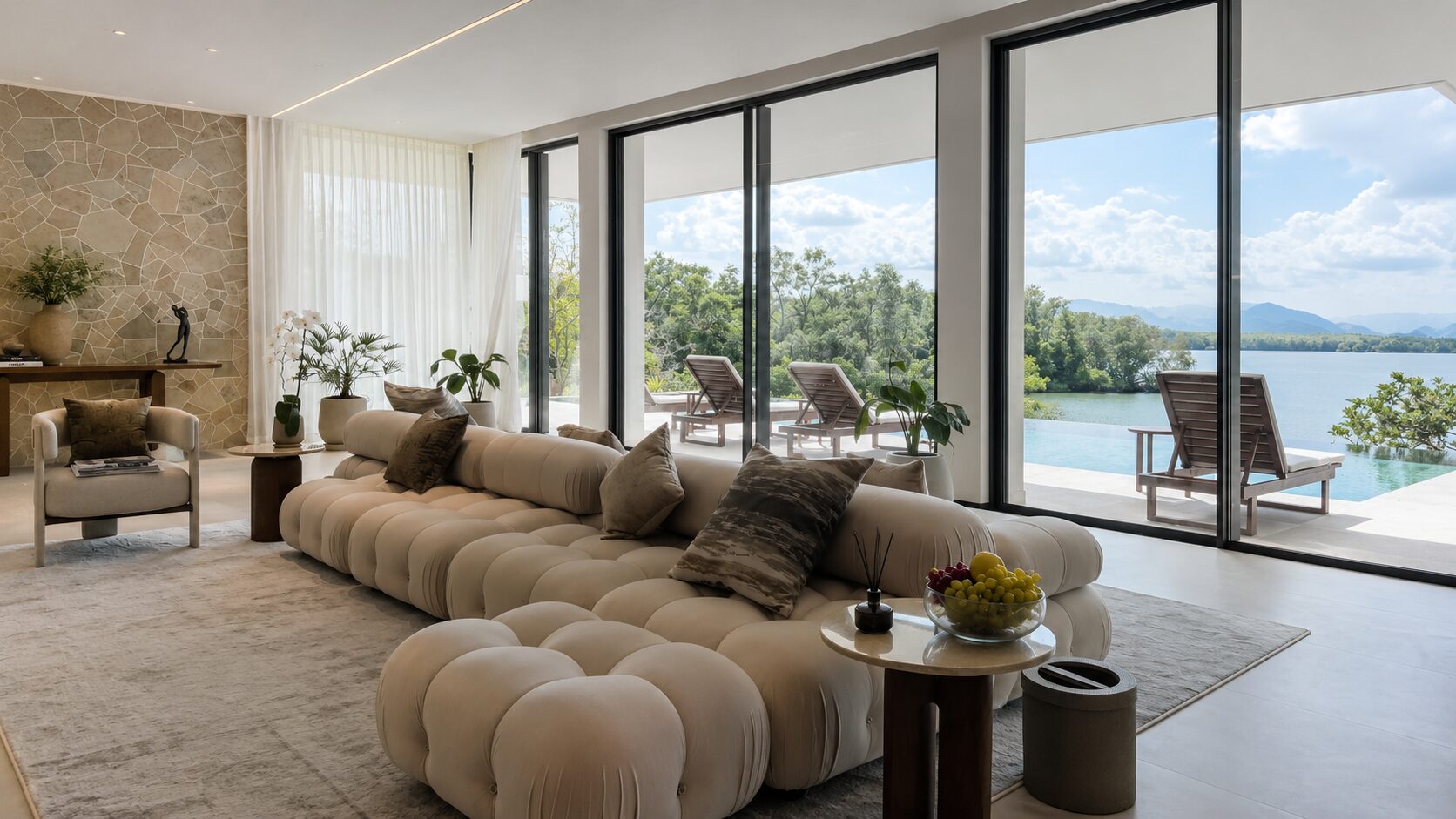 Luxurious living room with a cream tufted sectional, neutral throw pillows, and a stone accent wall, opening to a terrace with a lake view through large glass doors.