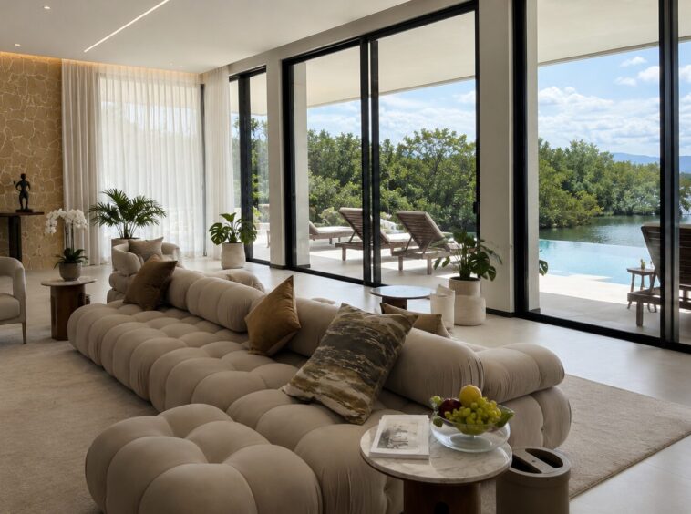 Luxurious living room with a long beige tufted sofa, neutral chairs, and potted plants, opening to a pool and lake view through floor-to-ceiling glass doors in a bright space.