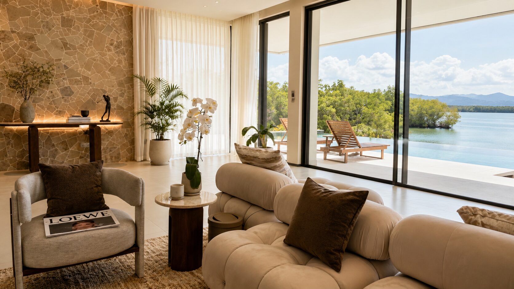 Luxurious living room with a cream tufted sofa and gray armchair, stone accent wall, plants, and floor-to-ceiling glass doors opening to a lake view.