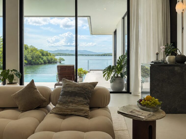 Luxurious living room with cream tufted sofa and cushions facing floor-to-ceiling glass doors opening to a lake view with trees and mountains in the distance.