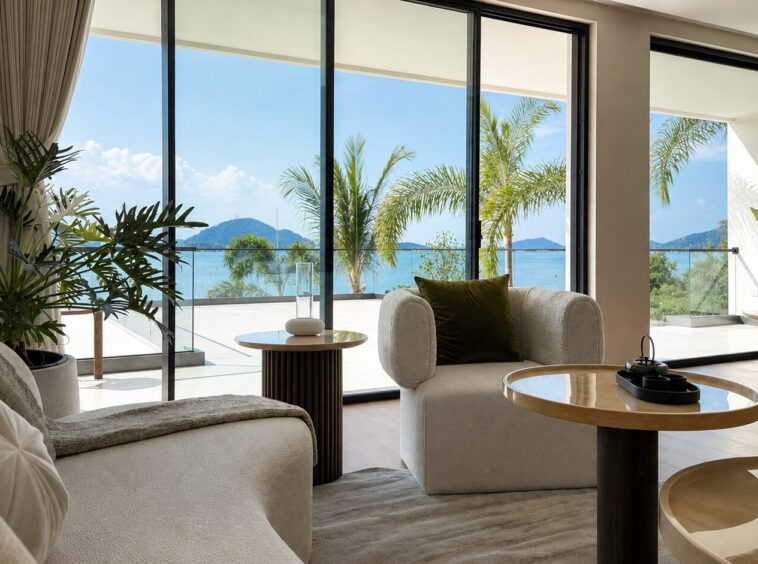 Bright living room with floor-to-ceiling glass doors opening to a sea view, palm trees outside, and beige sofas with a green cushion.