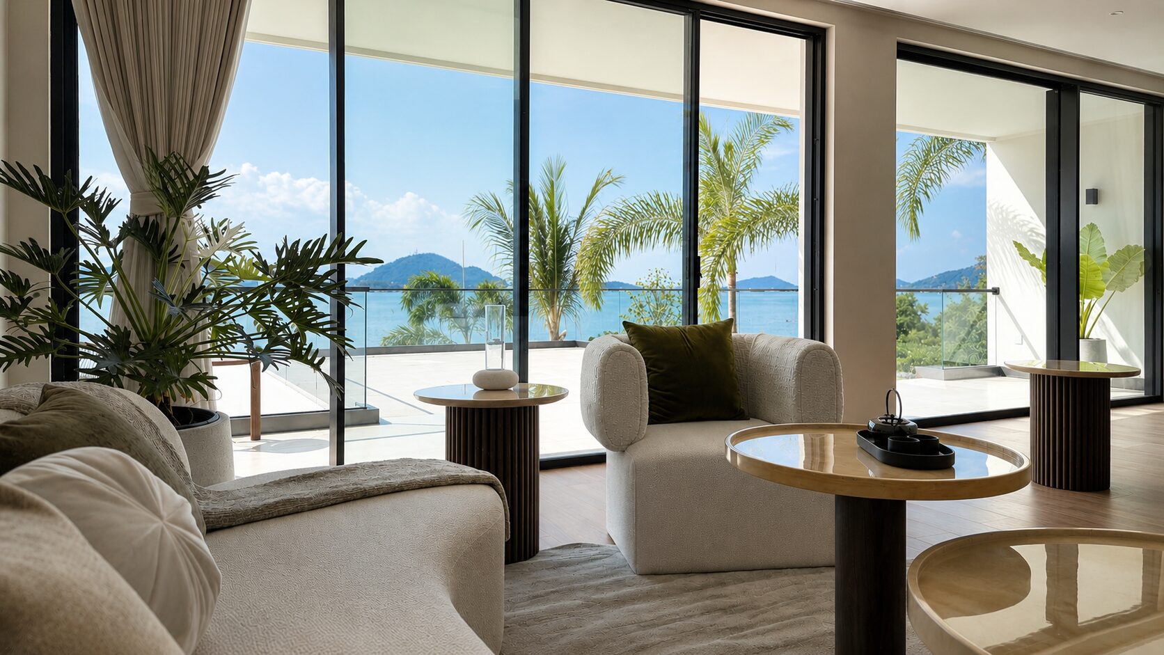 Bright living room with floor-to-ceiling glass doors opening to a sea view, palm trees outside, and beige sofas with a green cushion.