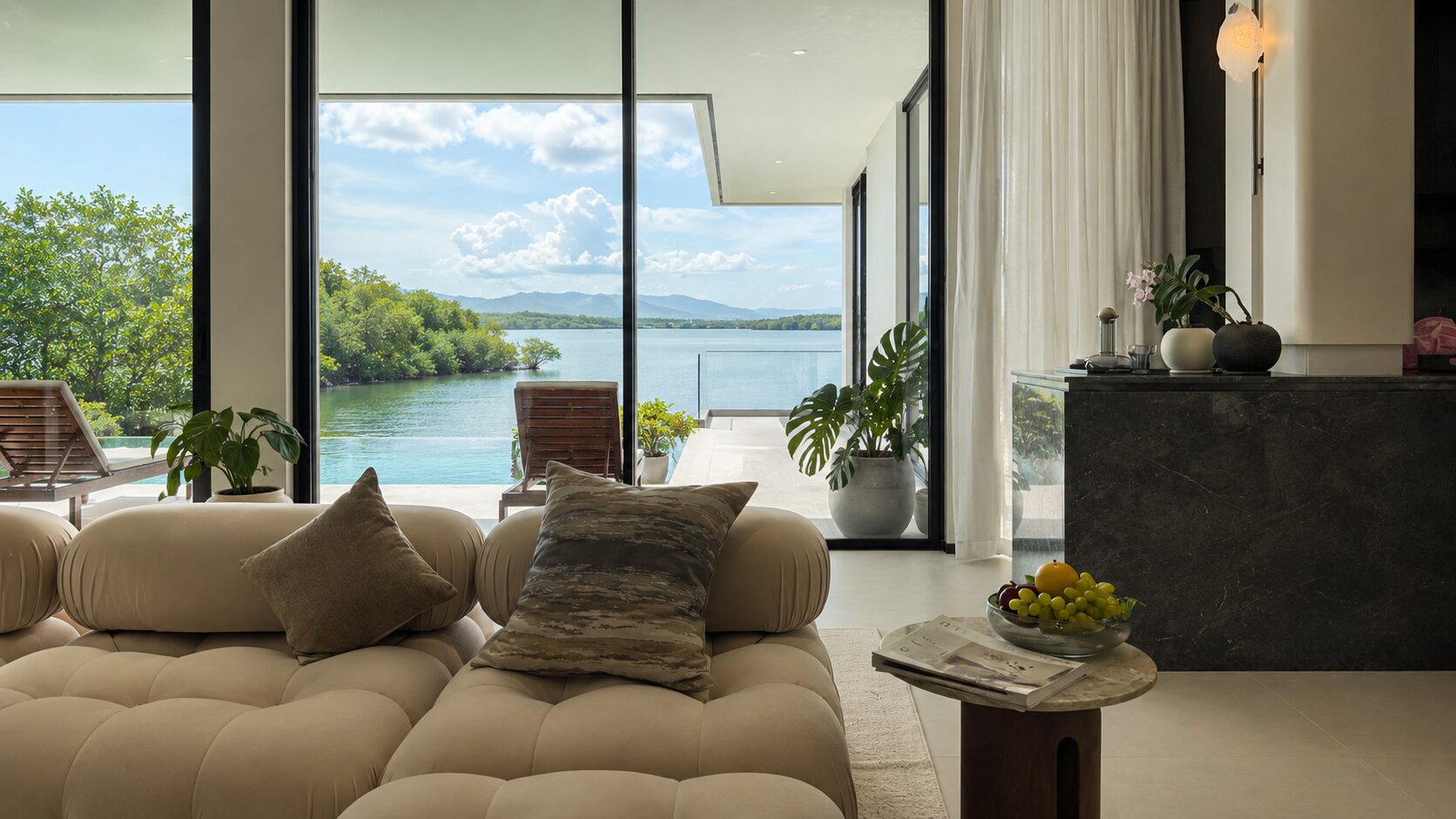 Luxurious living room with cream tufted sofa and cushions facing floor-to-ceiling glass doors opening to a lake view with trees and mountains in the distance.