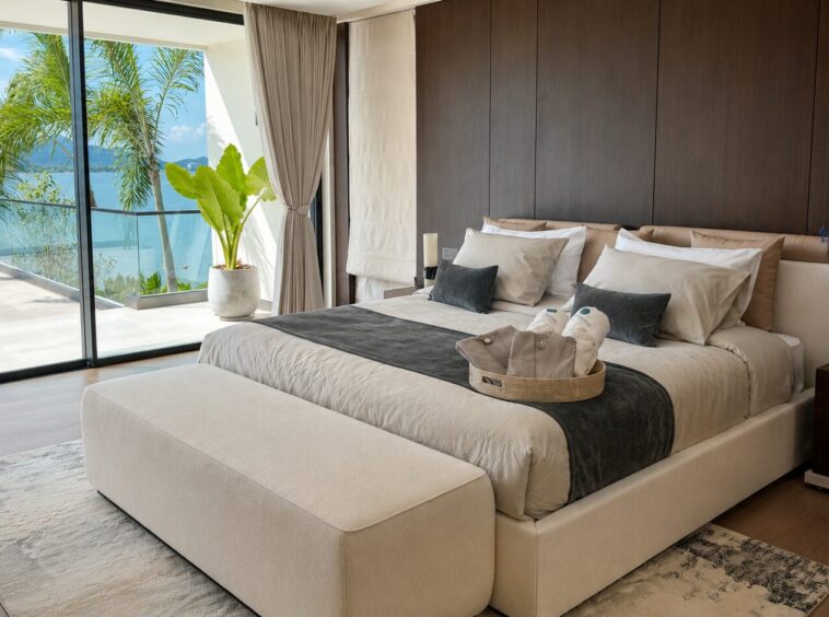 Luxurious bedroom with a king bed, beige upholstery, and a balcony view of palm trees and the sea.