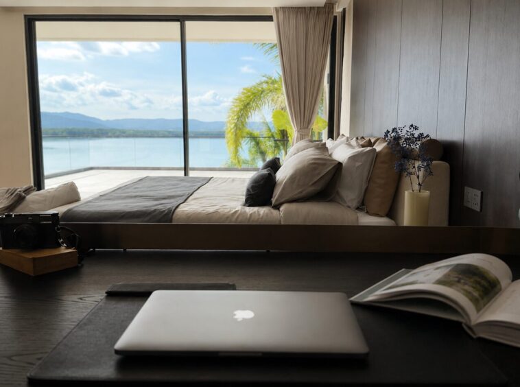 A modern bedroom with a desk and a laptop facing a bed and large glass doors opening to a lake view.
