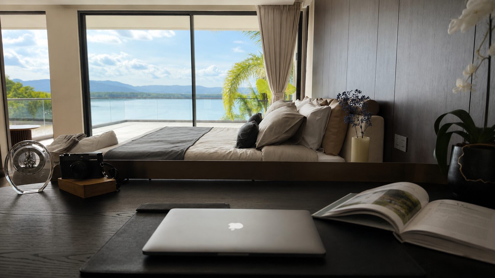 A modern bedroom with a desk and a laptop facing a bed and large glass doors opening to a lake view.