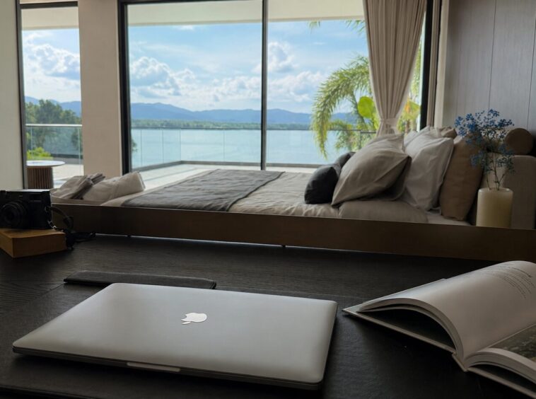 Work desk in a stylish hotel room with a laptop, open magazine, camera, and a glass clock, overlooking a balcony and water beyond.