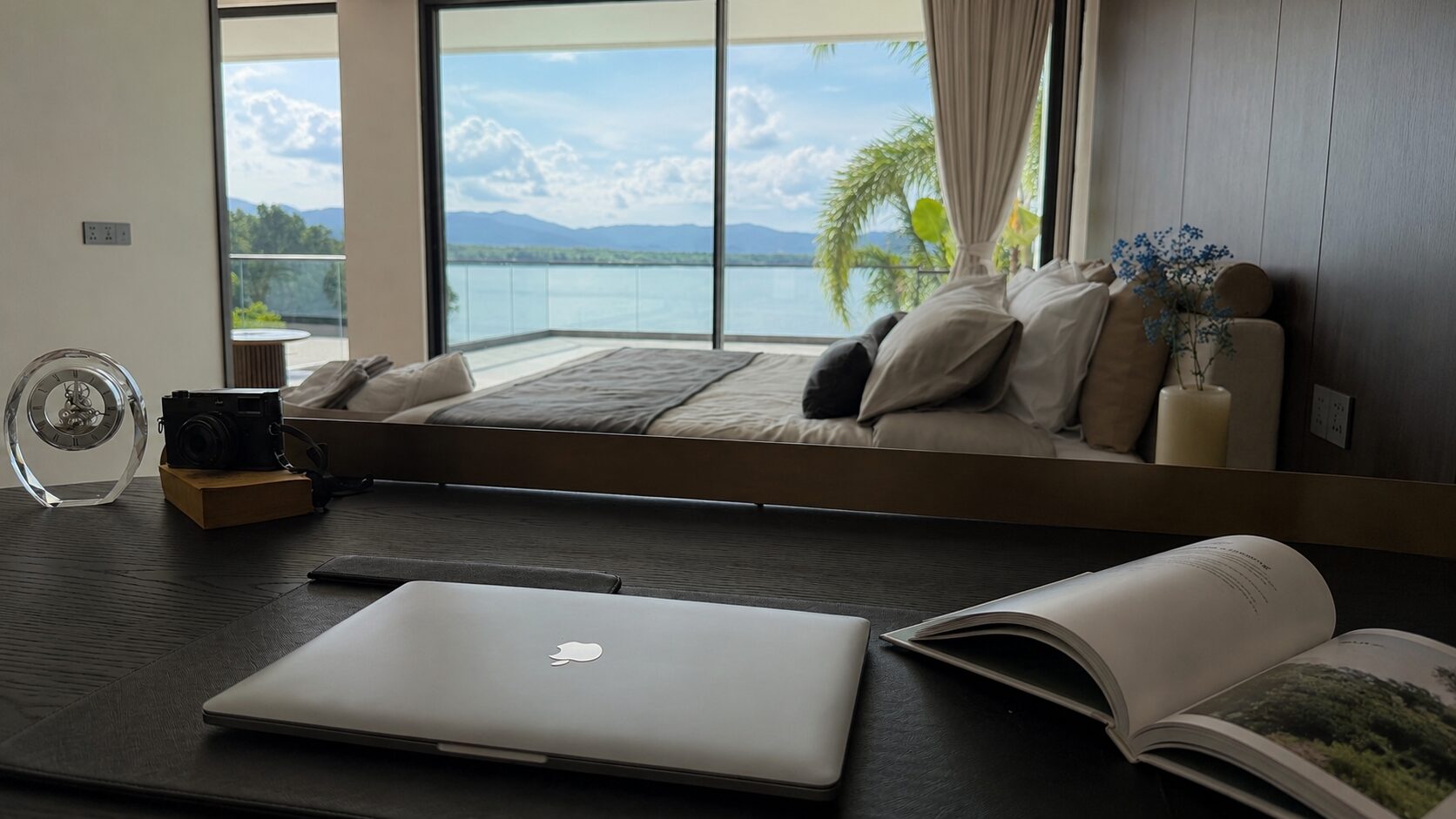 Work desk in a stylish hotel room with a laptop, open magazine, camera, and a glass clock, overlooking a balcony and water beyond.