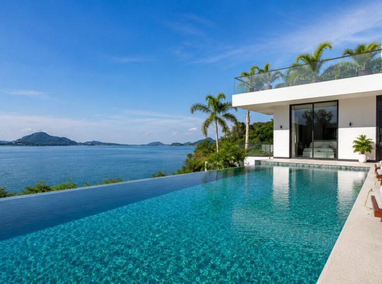 Infinity pool at a modern villa with an ocean view and palm trees under a blue sky, lounge chairs nearby.
