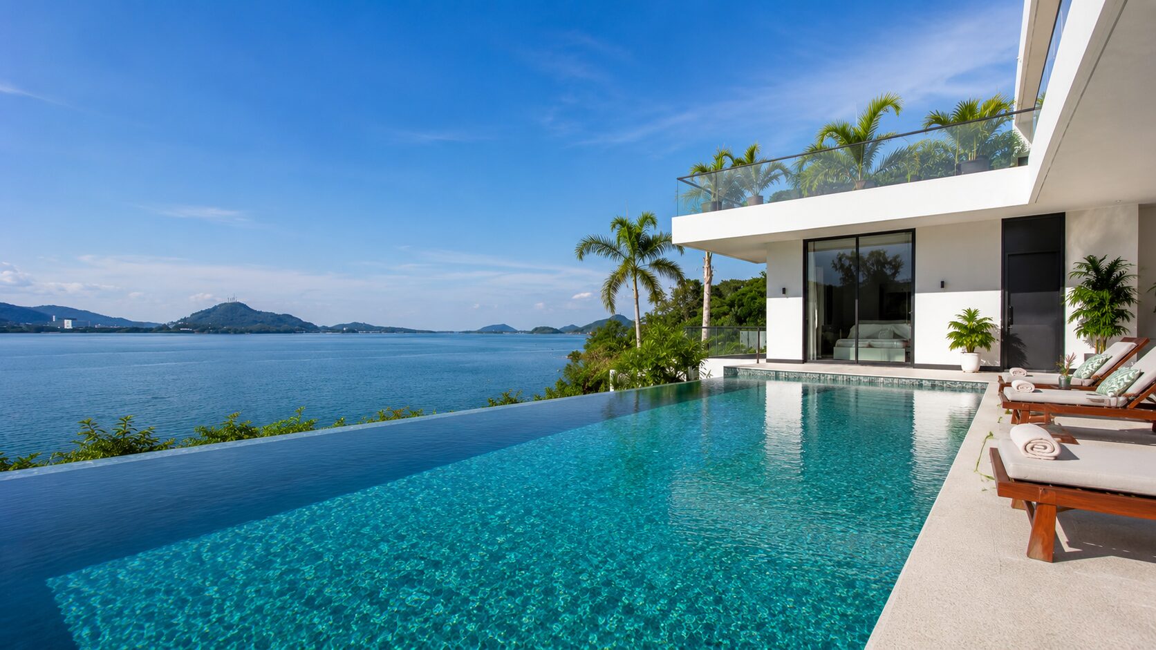 Infinity pool at a modern villa with an ocean view and palm trees under a blue sky, lounge chairs nearby.