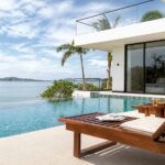 Modern white villa with glass balcony overlooking the ocean, palm trees and a poolside wooden lounge chair in the foreground.