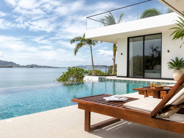 Modern white villa with glass balcony overlooking the ocean, palm trees and a poolside wooden lounge chair in the foreground.