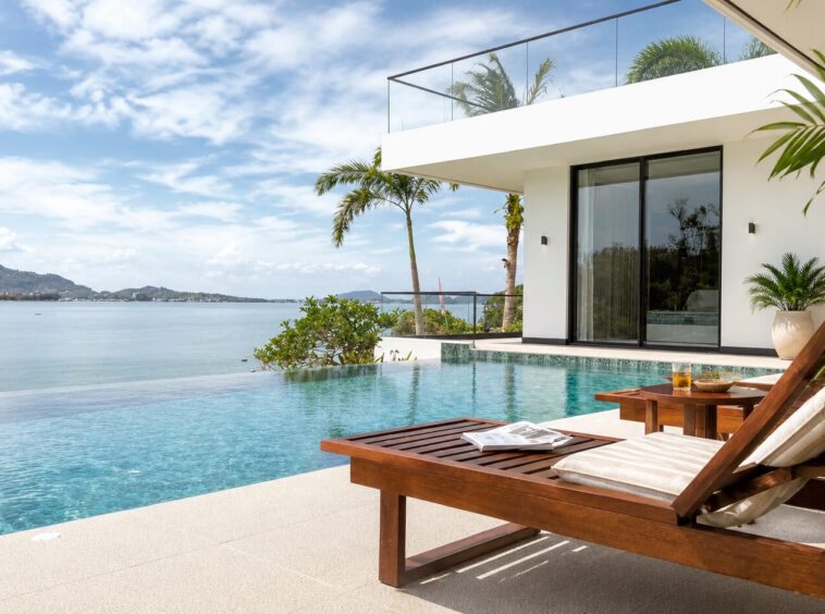 Modern white villa with glass balcony overlooking the ocean, palm trees and a poolside wooden lounge chair in the foreground.
