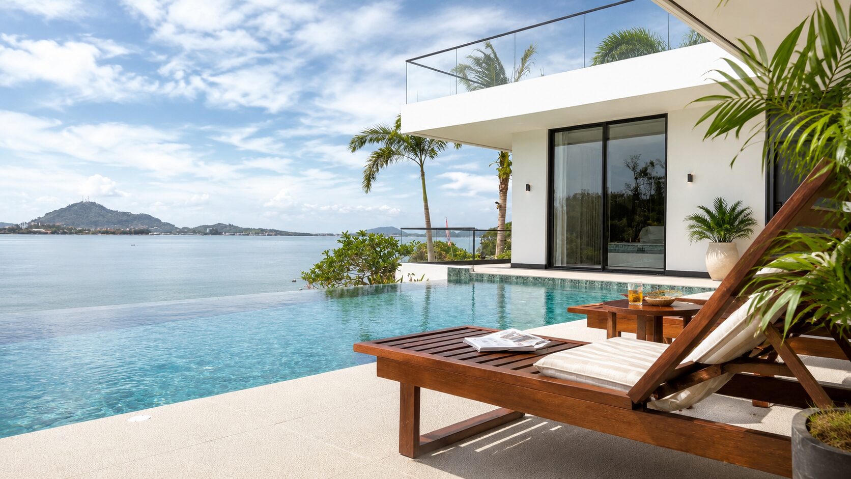 Modern white villa with glass balcony overlooking the ocean, palm trees and a poolside wooden lounge chair in the foreground.